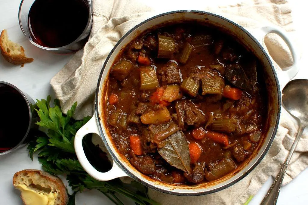 A rustic Dutch oven venison stew simmering with chunks of tender venison, carrots, celery, and bay leaves in a rich, deep brown broth. Surrounding the pot are glasses of red wine, fresh parsley, and crusty bread with butter, creating a cozy and inviting meal setting.
